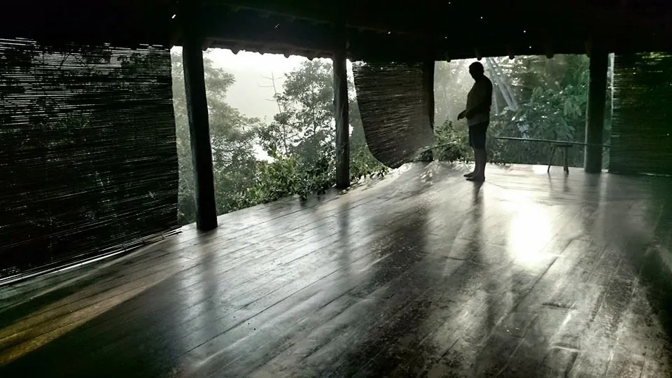 The silhouette of a person standing on the wooden floor of an open hut, with mist outside.