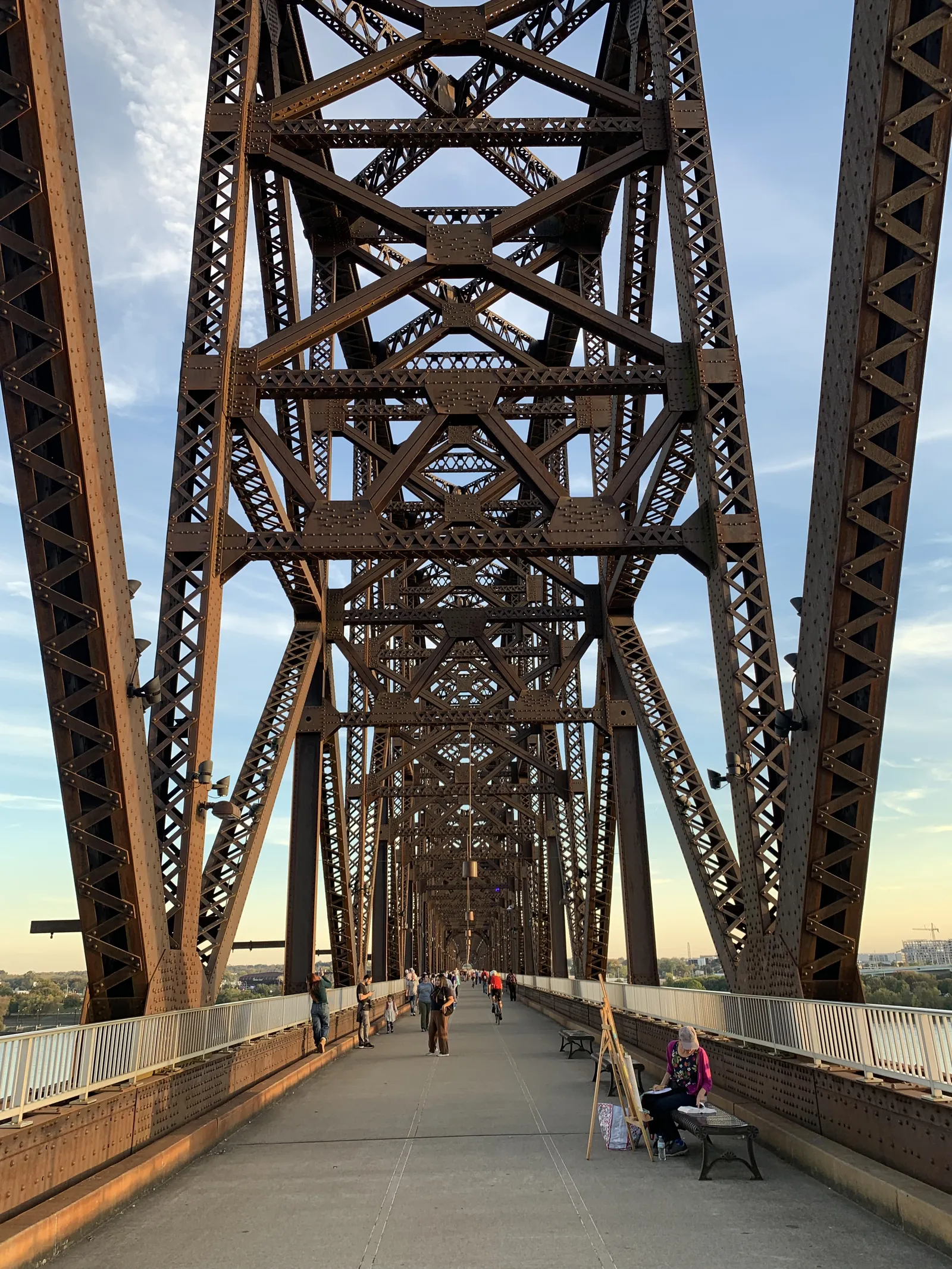 People walking across a rust colored steel truss bridge at sunset, with a woman on a bench painting at an easel on the right