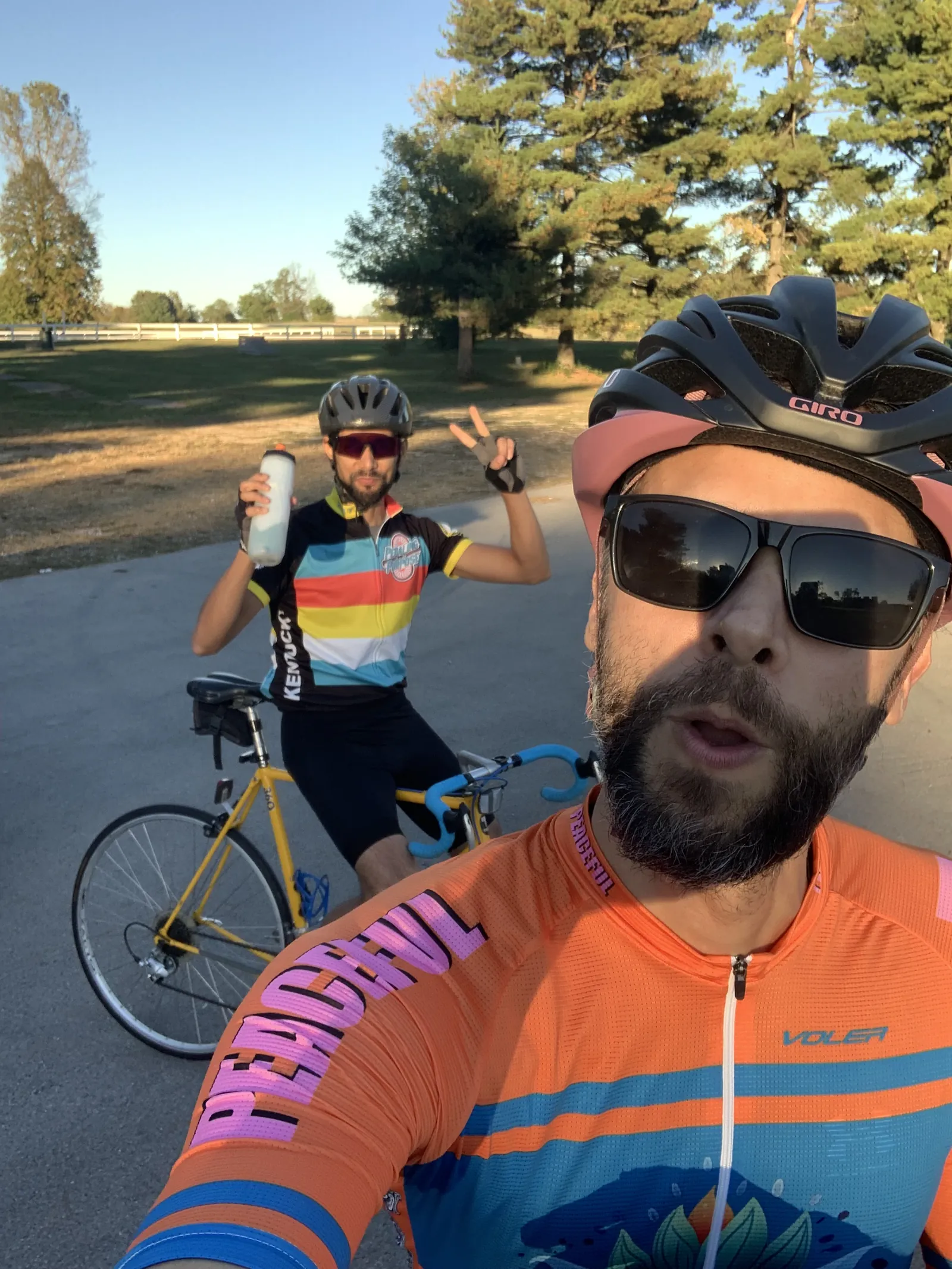 Selfie of Diego in a helmet, sunglasses and an orange jersey on a paved road, with a friend behind him on a yellow road bike holding up a water bottle and a peace sign