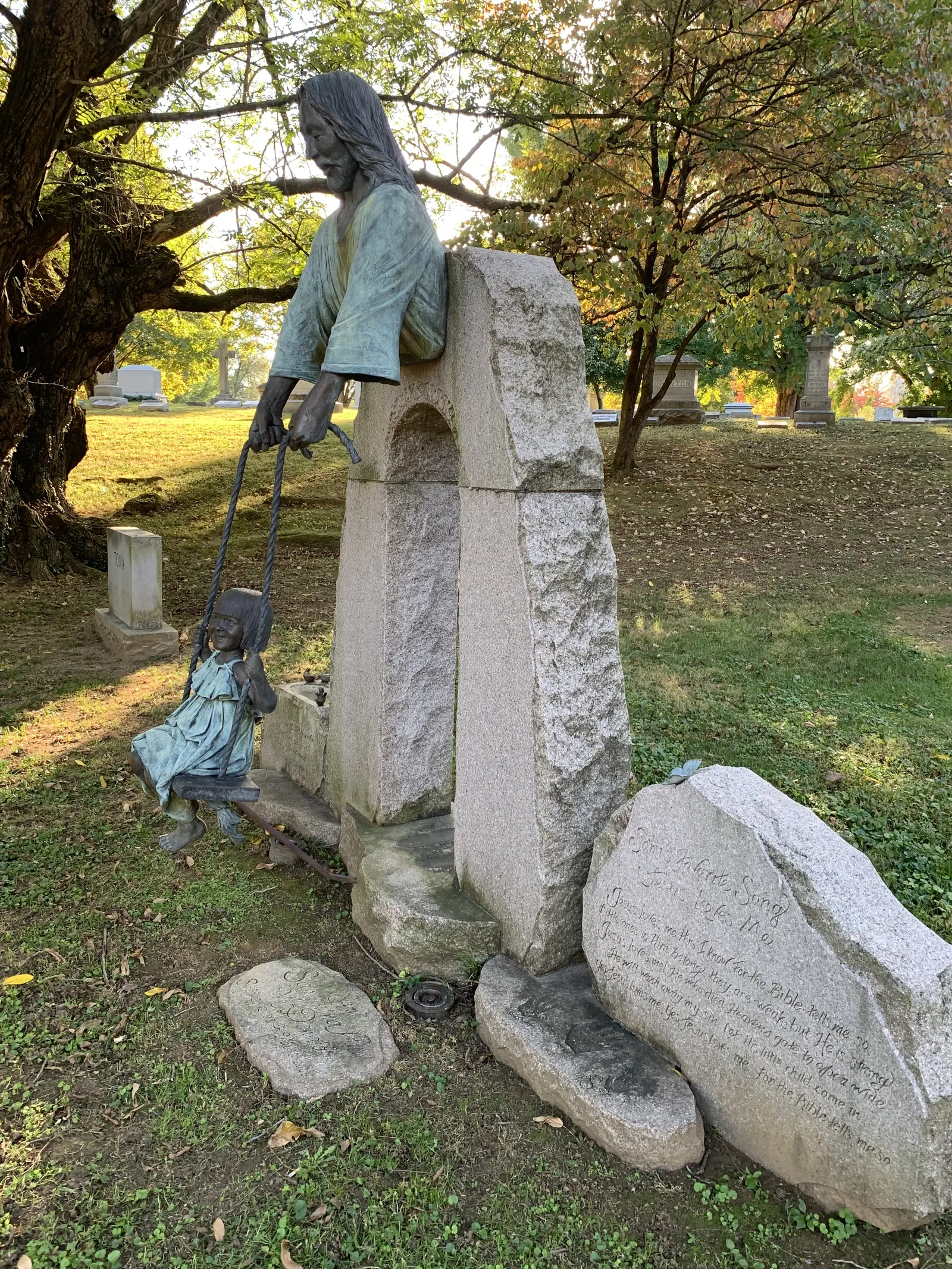 Cemetery monument with a bronze Jesus leaning over a granite block and holding the ropes of a swing, with a bronze little girl sitting on it, under autumn trees