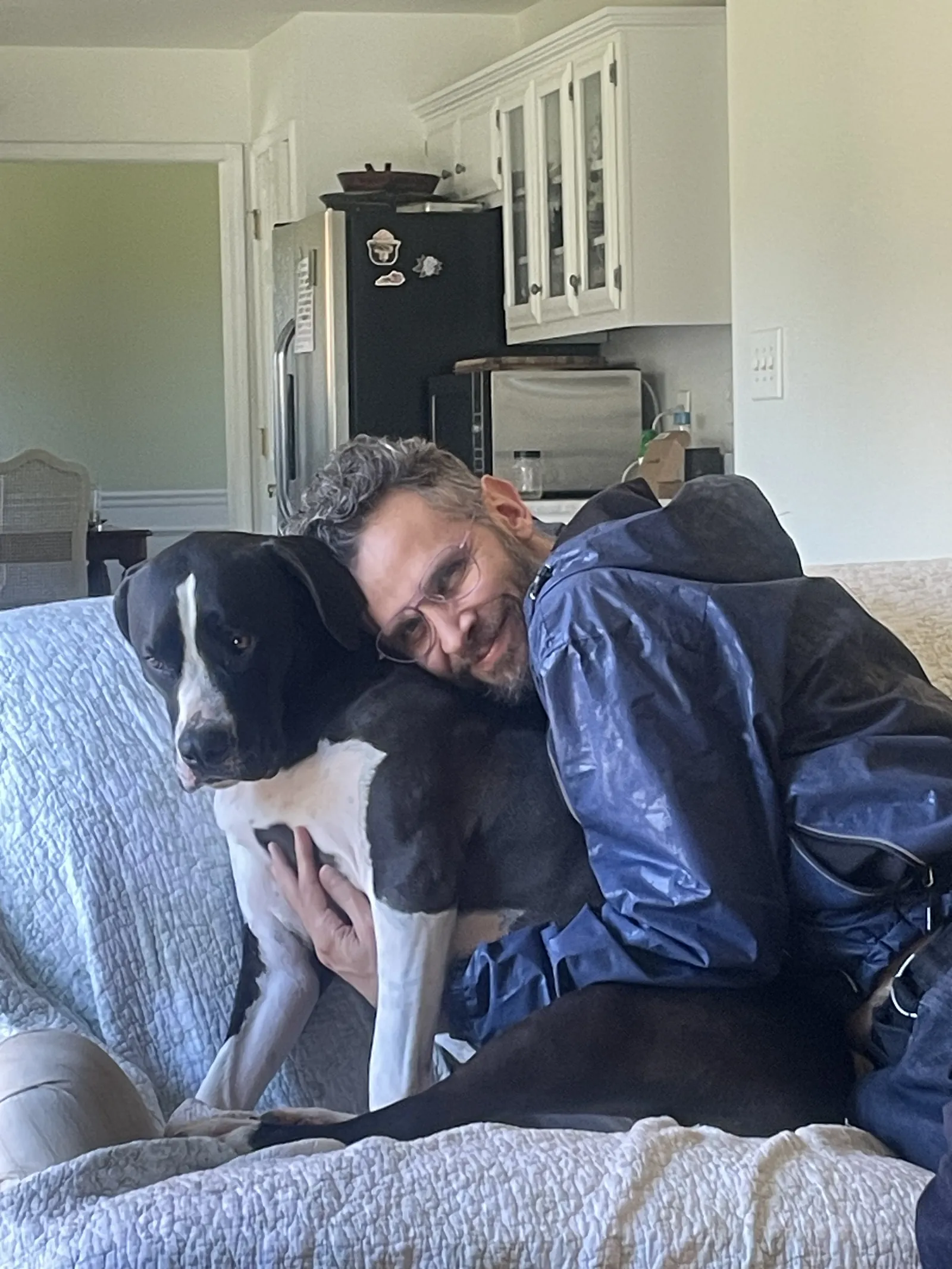 Diego in a blue rain jacket and round glasses hugging Mapple, a big black and white dog, on a couch with a kitchen behind them