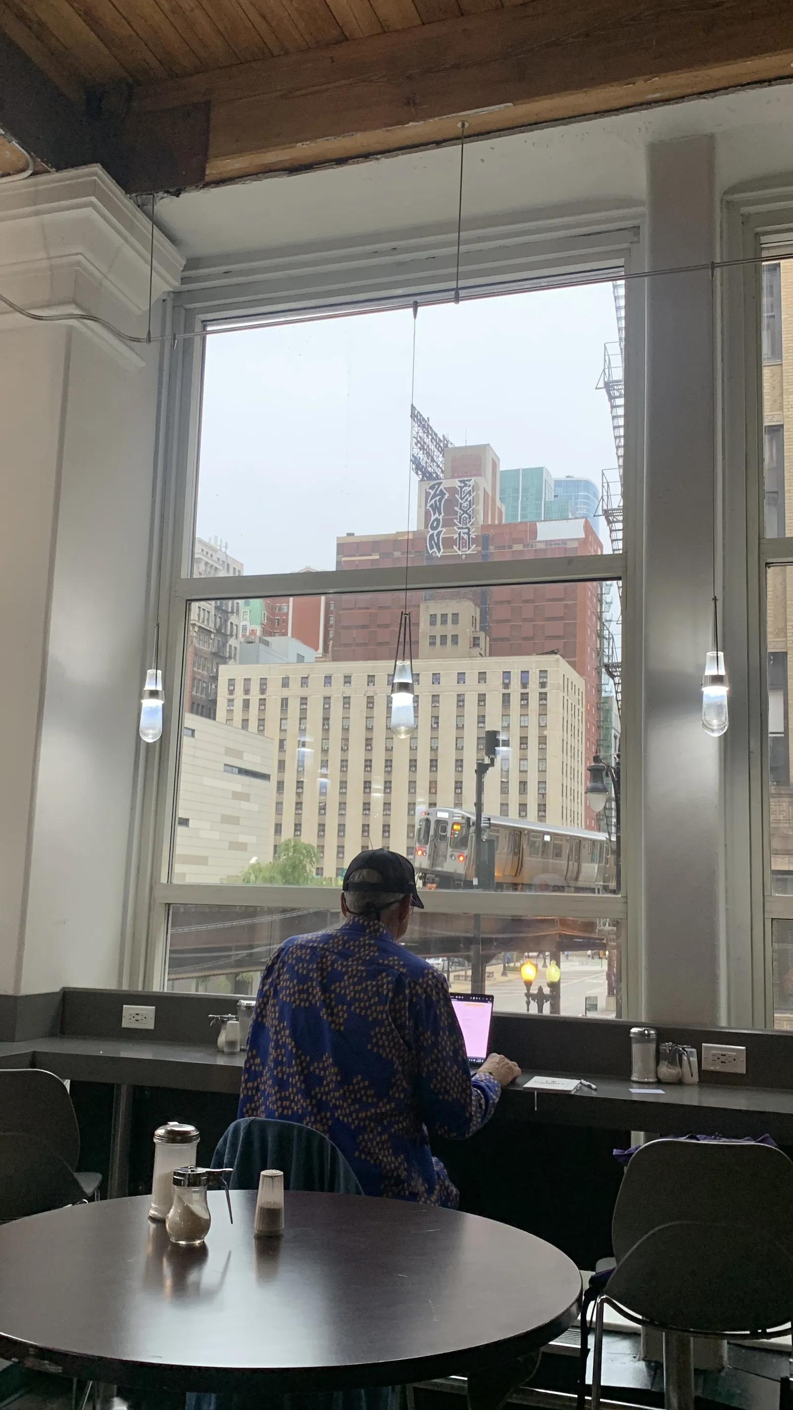 A man in a cap and a patterned blue shirt working on a laptop at a café window, with an elevated L train passing between old brick buildings outside