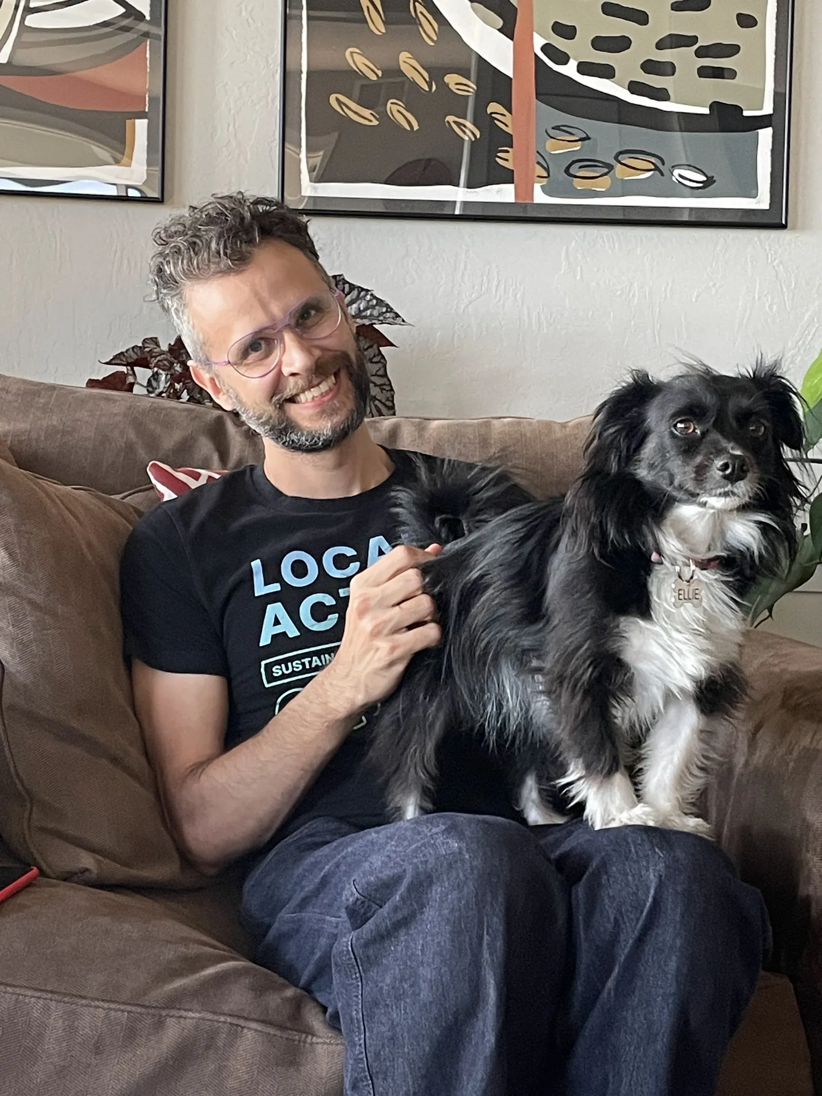 Diego smiling on a brown couch in purple glasses, holding Ellie, a small black and white long haired dog with a name tag, who looks away from the camera