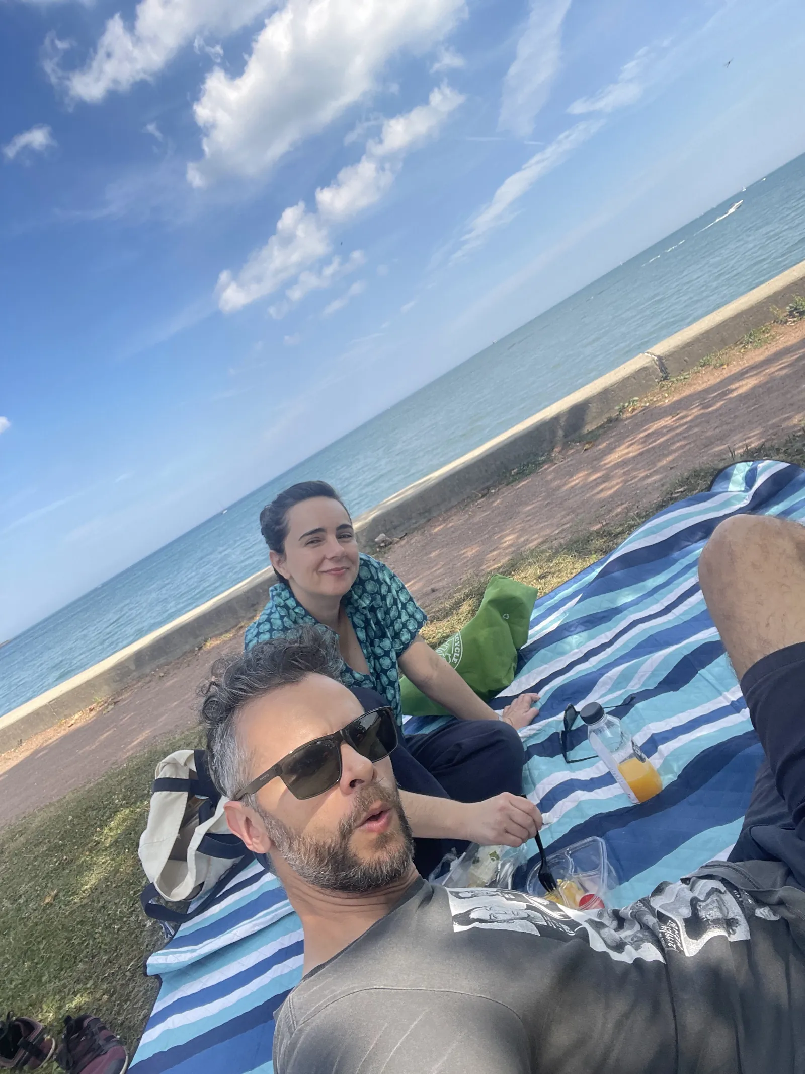 Tilted selfie of Diego in sunglasses lying on a striped picnic blanket with Leandra sitting behind him, Lake Michigan and a blue sky in the background